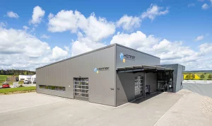 Außenansicht des Firmengebäudes der Hammer Automationstechnik GmbH in Egenhausen, Anbieter von Handschraubern, EC Schrauber, Schraubtechnik, Schraub- und Setzeinheiten, Zuführtechnik sowie Drehmomentmessgeräten für Deutschland, Österreich und die Schweiz. Das Gebäude erscheint in grau. Es ist jeweils auf der linken und der Frontseite des Gebäudes ein Rolltor zu sehen. Hier ist außerdem der Schriftzug "Hammer Automationstechnik" zu sehen.